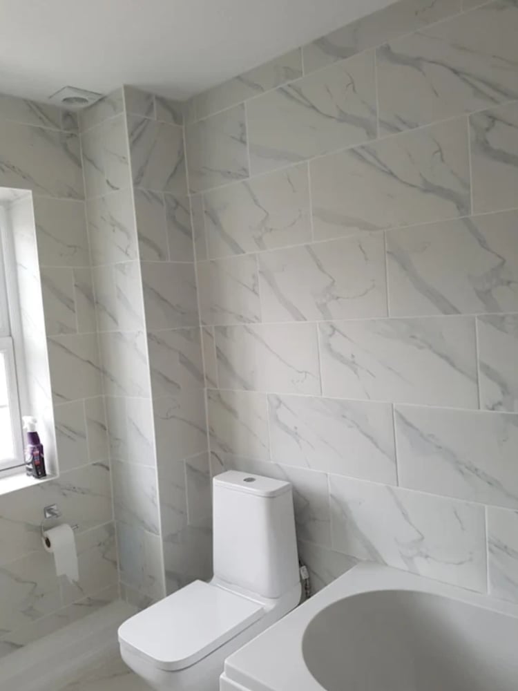 Modern bathroom with white marble-look wall tiles, toilet, round sink, and small window with light fixture