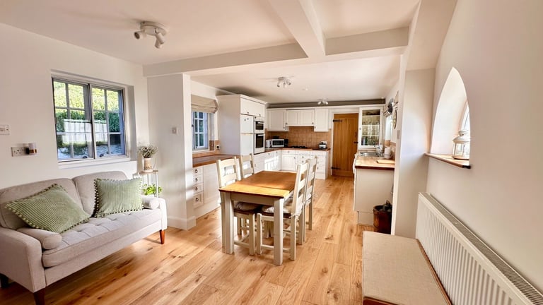 Open-plan kitchen and living room with cream and wood tones, featuring a breakfast bar, sofa, and natural light from large windows