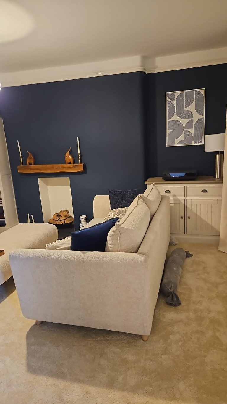 Modern living room with cream armchair against dark blue accent wall, wooden shelf, and white cabinet with geometric wall art