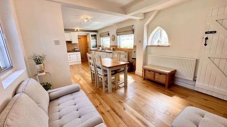Open-plan living and dining room with wooden flooring, cream sofa, dining table, and kitchen visible in background