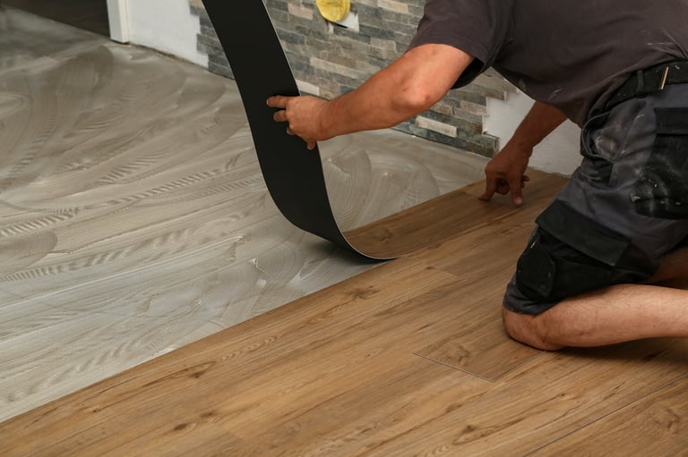 Person kneeling on wooden floor while installing vinyl flooring with hands smoothing down a black edge piece