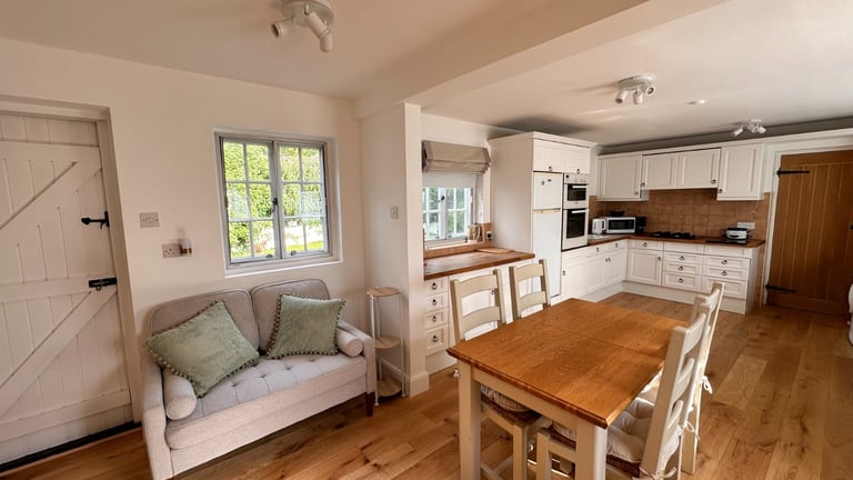 Open-plan kitchen-dining area with wooden table, white cabinetry, window seat with cushions, and hardwood flooring
